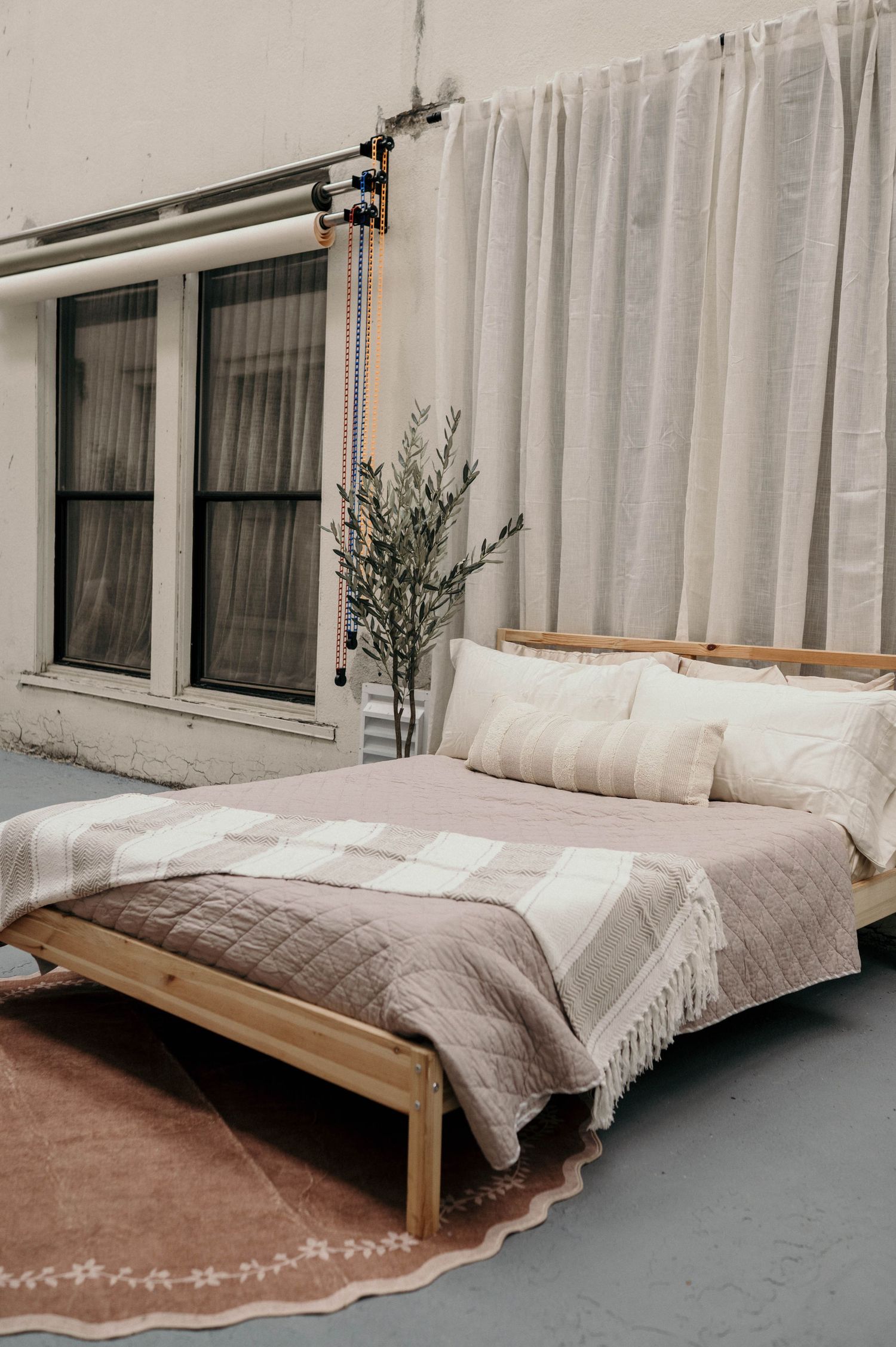 Minimalist bedroom with wooden bed frame, neutral bedding, fringed blanket, area rug, and sheer curtains by industrial windows.
