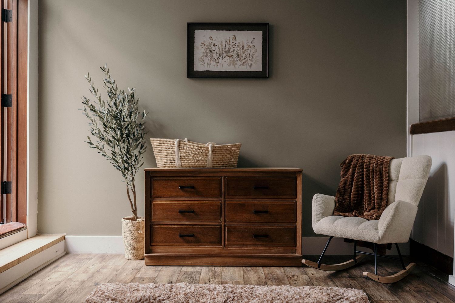 Cozy bedroom corner with walnut dresser, white rocking chair, olive tree, and framed art on sage green wall.