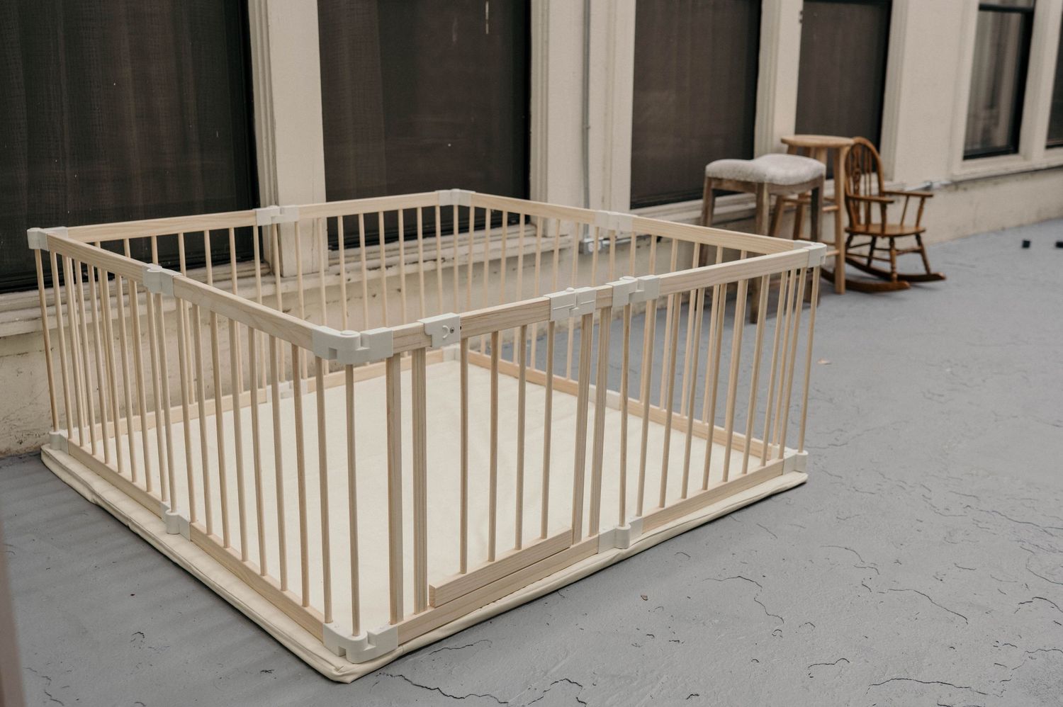 Large wooden baby playpen with vertical slats on gray floor, with rocking chair in background.