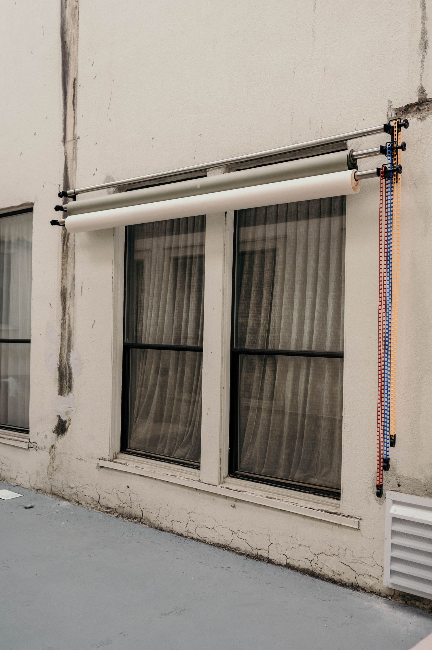 Exterior wall with two large windows, sheer curtains, and colorful pipe installations on a weathered building facade.