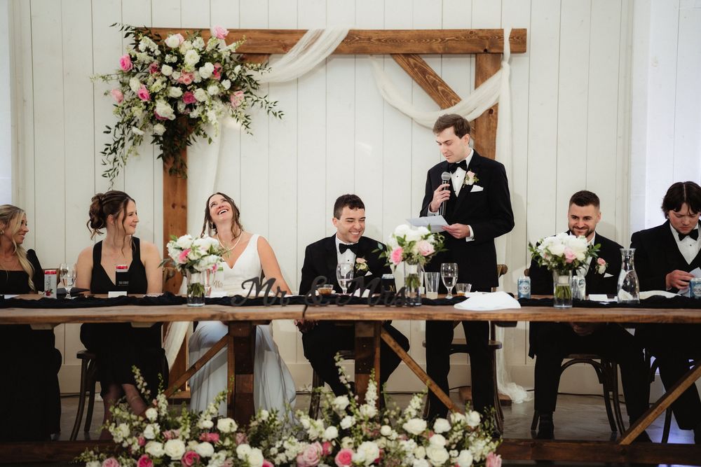 Wedding reception head table with bride and groom seated as groomsman delivers a heartfelt speech in a floral barn venue.