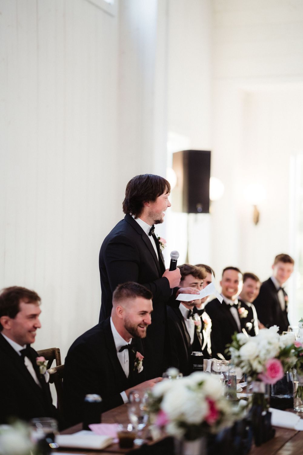 Groomsman delivers a heartfelt wedding speech at the head table while guests in black tuxedos listen and smile.