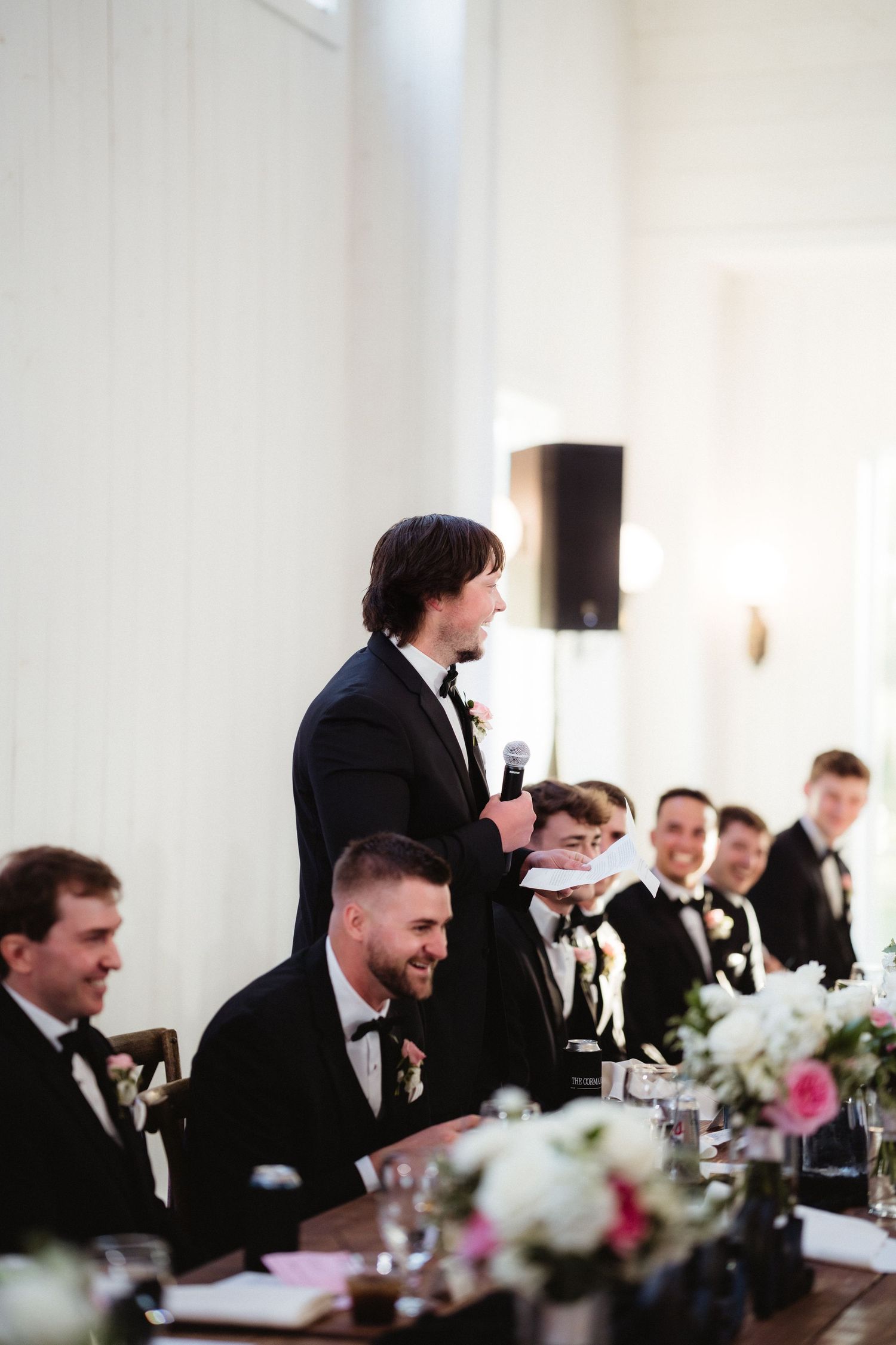 Groomsman delivers a heartfelt wedding speech at the head table while guests in black tuxedos listen and smile.
