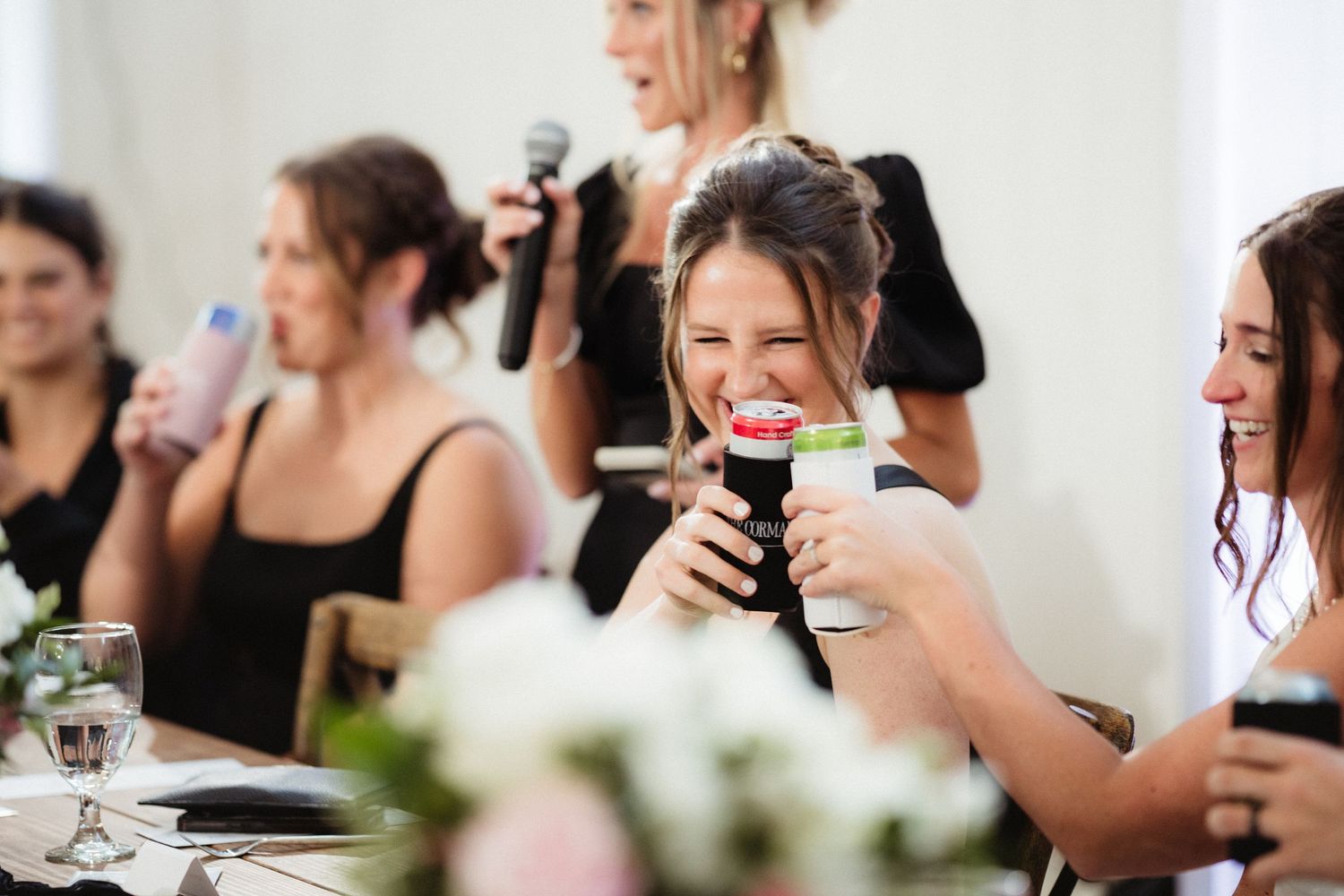 Women in black dresses laughing and holding drinks at a wedding reception celebration.