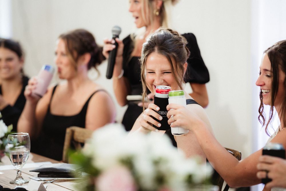 Women in black dresses laughing and holding drinks at a wedding reception celebration.