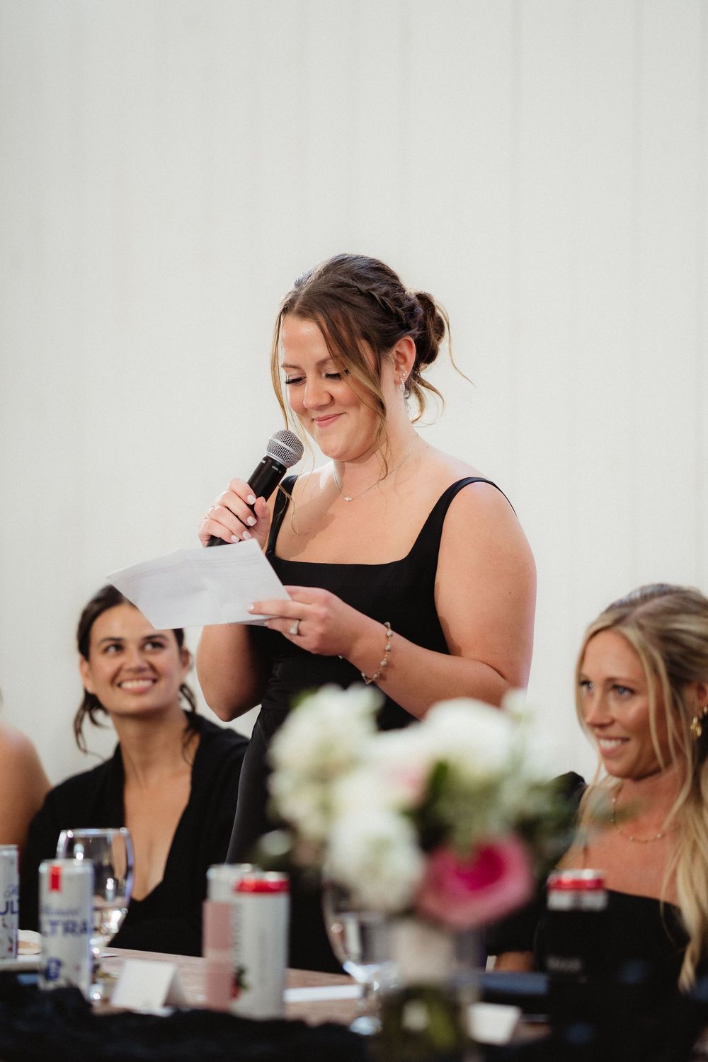 Woman in black dress giving a speech at a wedding reception, holding microphone and notes, smiling guests seated behind her.