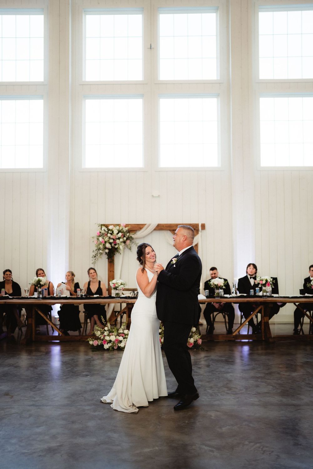 Bride and groom share their first dance in an elegant white barn wedding venue with large windows and guests seated at long tables.