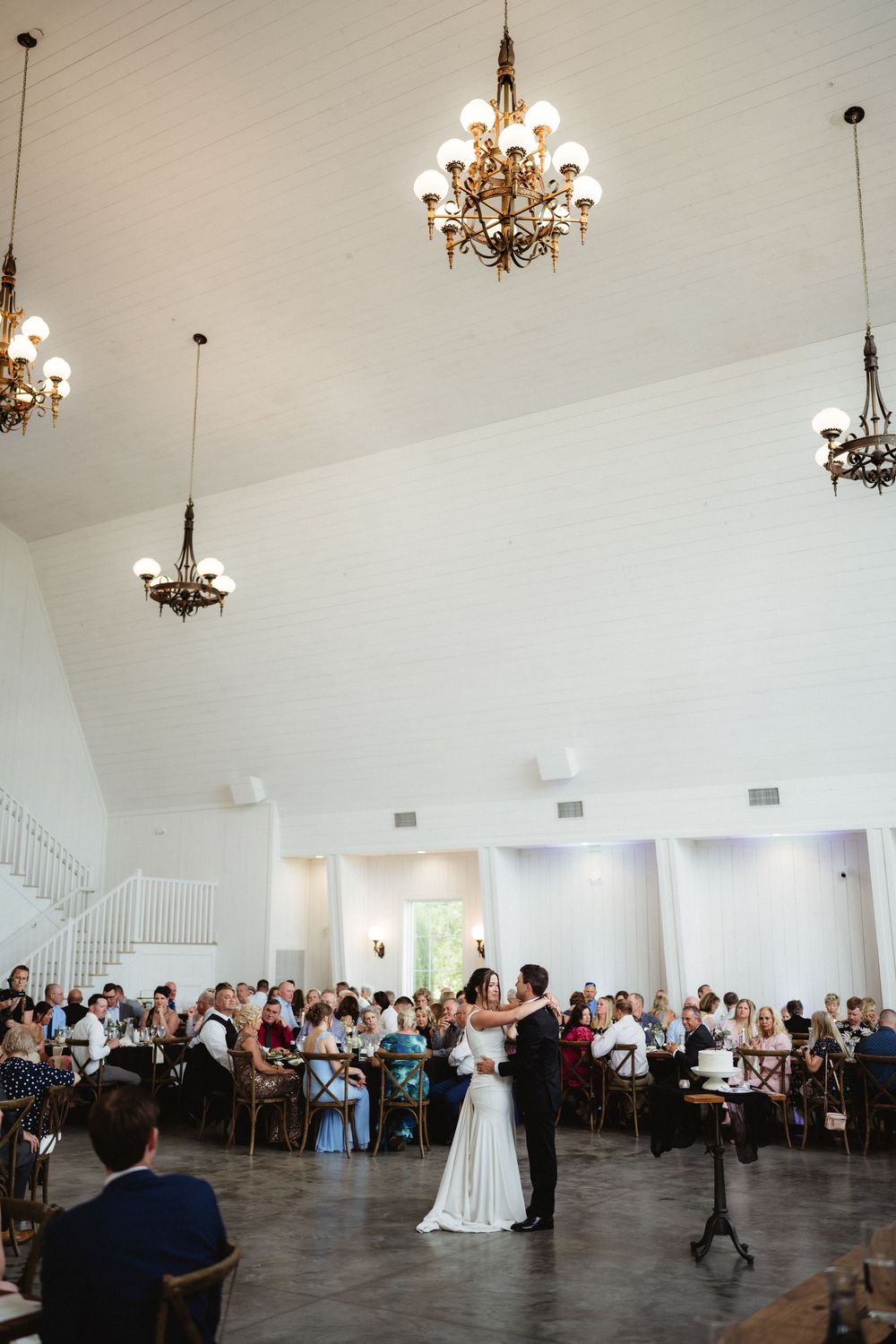 Bride and groom share their first dance in an elegant white venue with chandeliers and seated wedding guests.