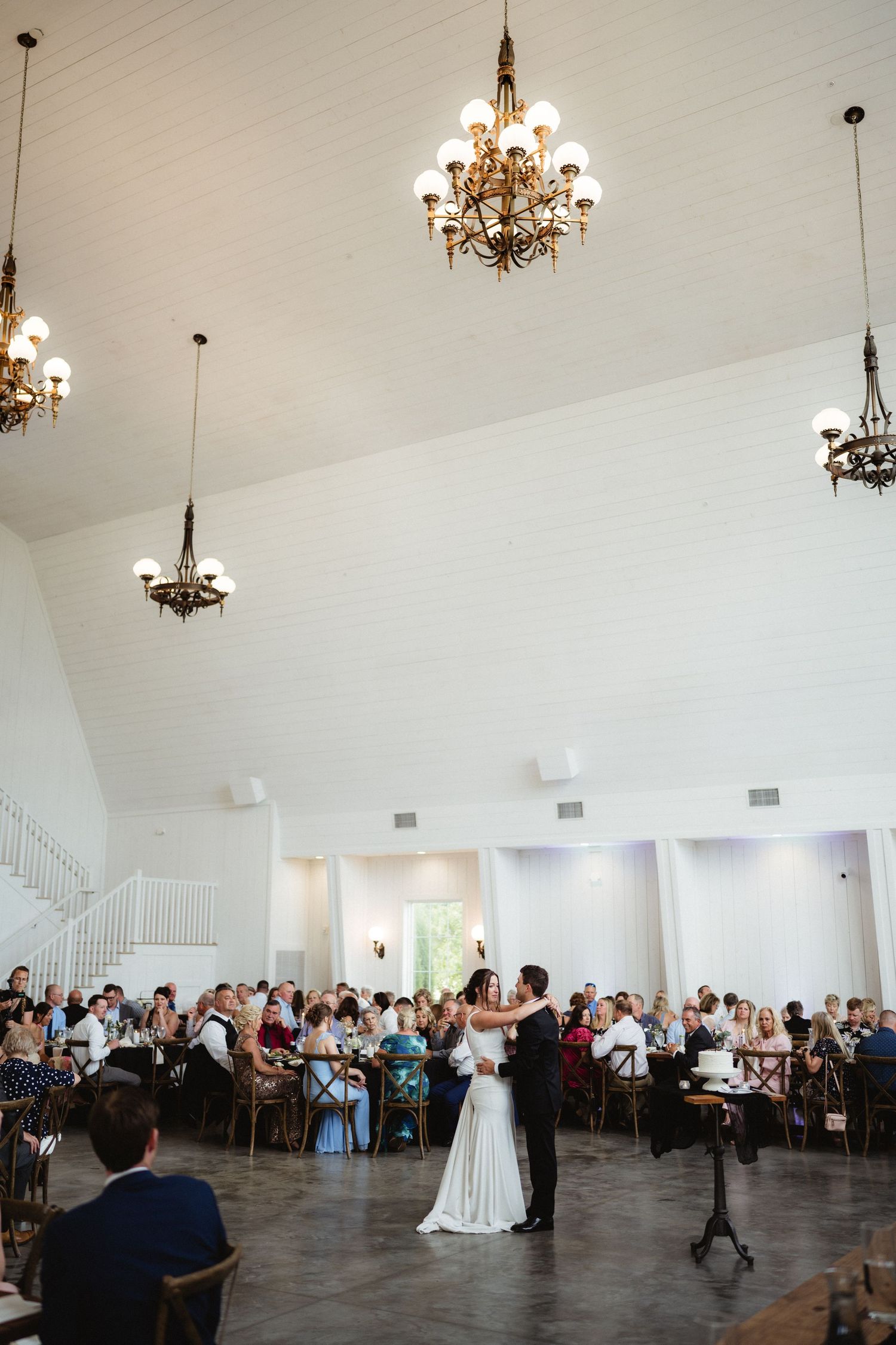 Bride and groom share their first dance in an elegant white venue with chandeliers and seated wedding guests.