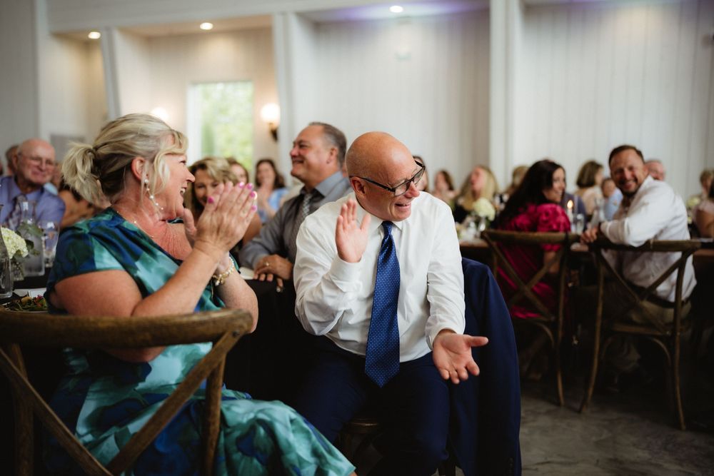 Wedding guest animatedly gestures while seated at a reception table as others clap and laugh around him.
