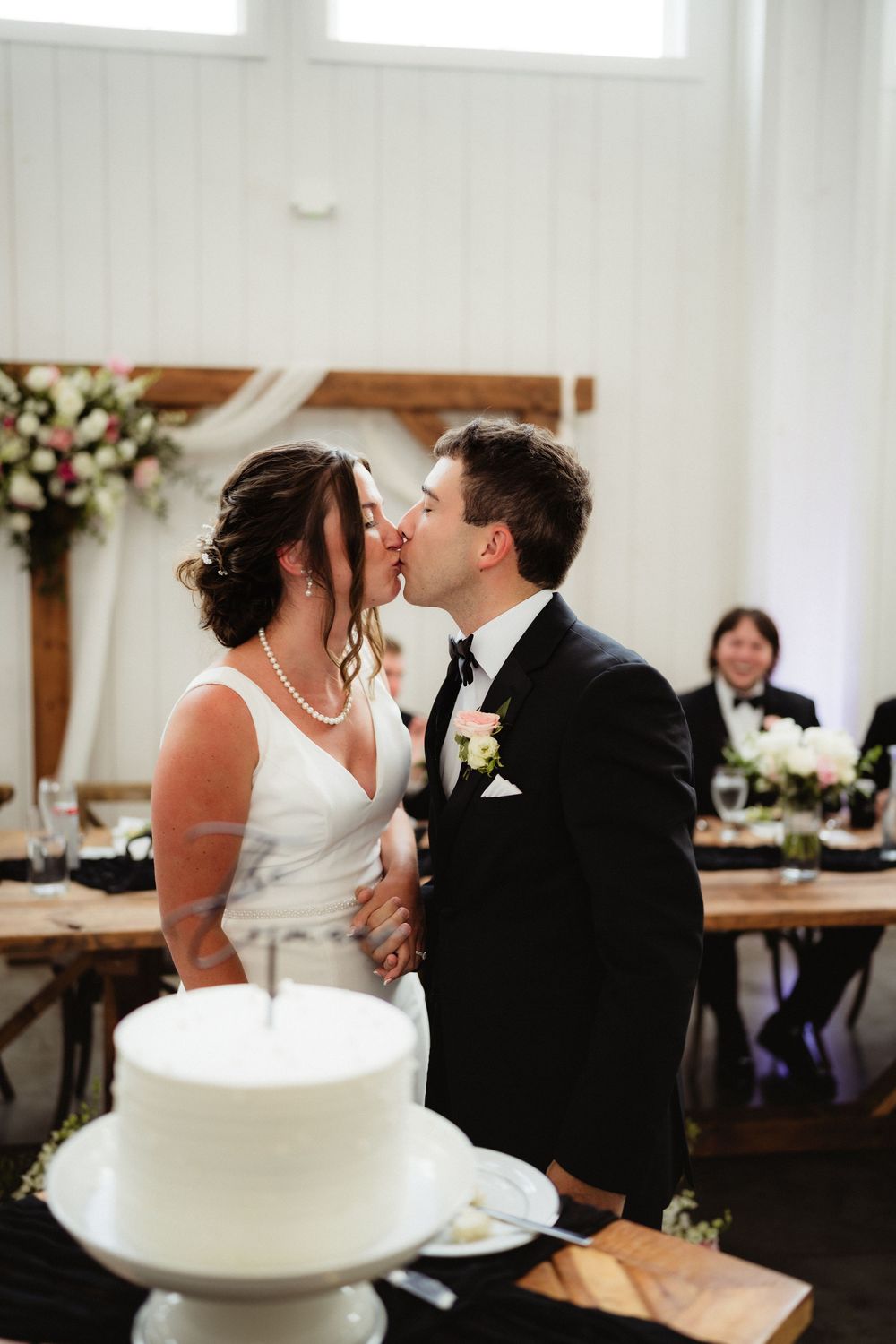 Bride and groom share a first kiss as a married couple beside their white wedding cake at their reception.