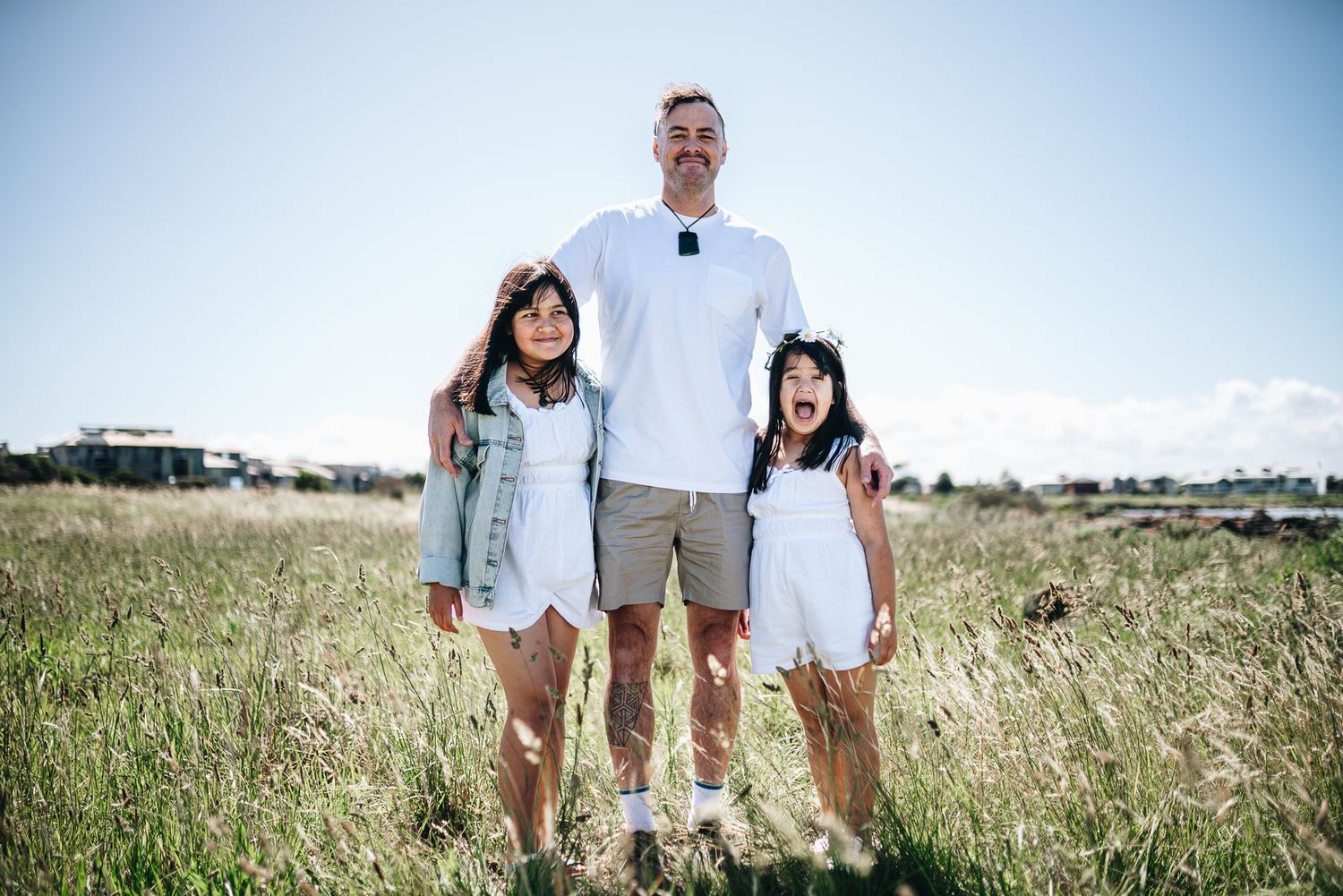 A father and two young daughters smile together in a sunny field, dressed in white and light colors for a family photo in Melbourne.