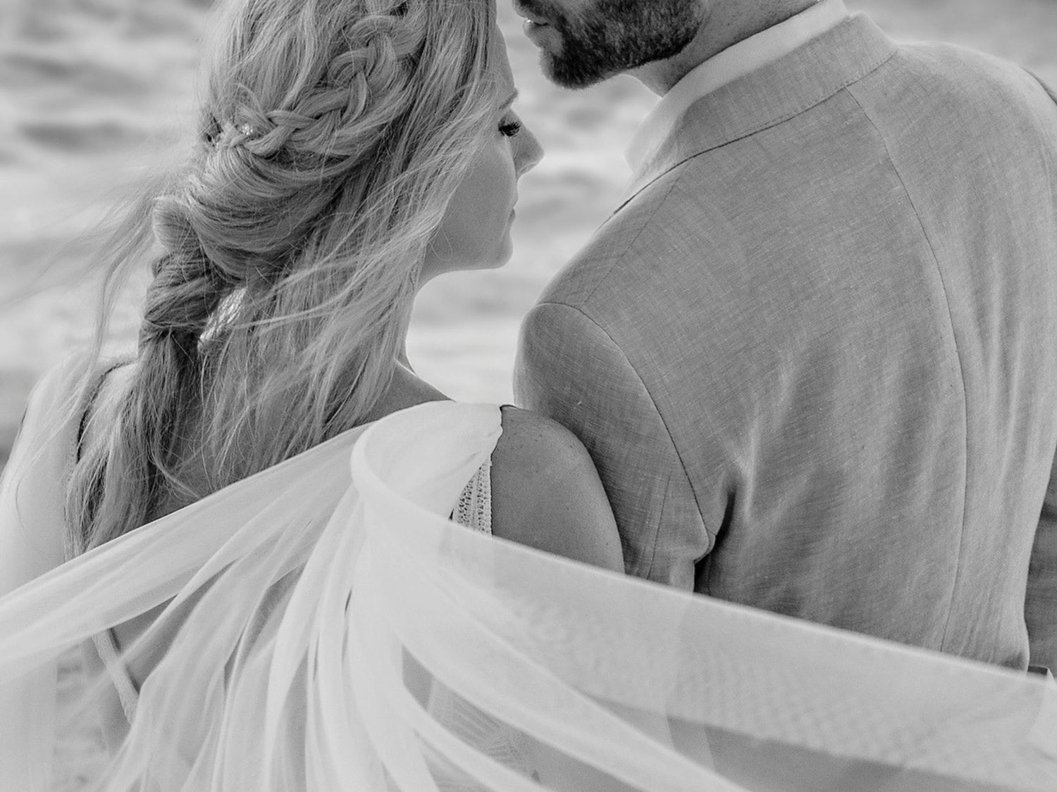 Black and white photo of a couple embracing on a beach, bride with braided hair and flowing veil.
