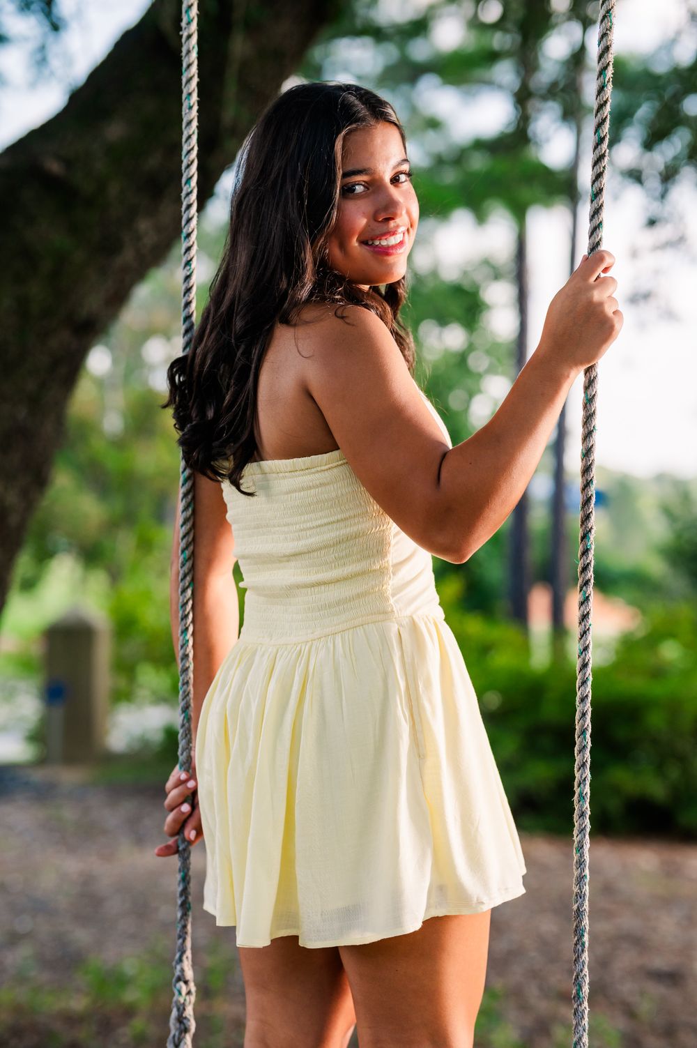 Smiling young woman in yellow strapless dress standing on a rope swing outdoors among green trees.
