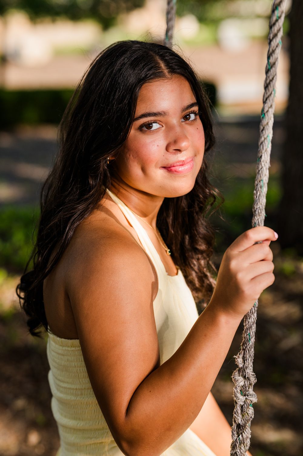 Young woman with long dark hair smiling while holding a rope swing outdoors in warm sunlight.