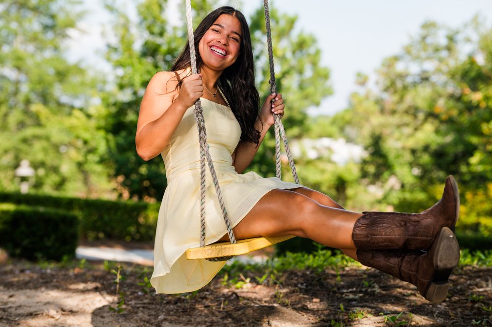 Young woman in yellow dress and brown cowboy boots smiling on a wooden swing in a sunny green park.