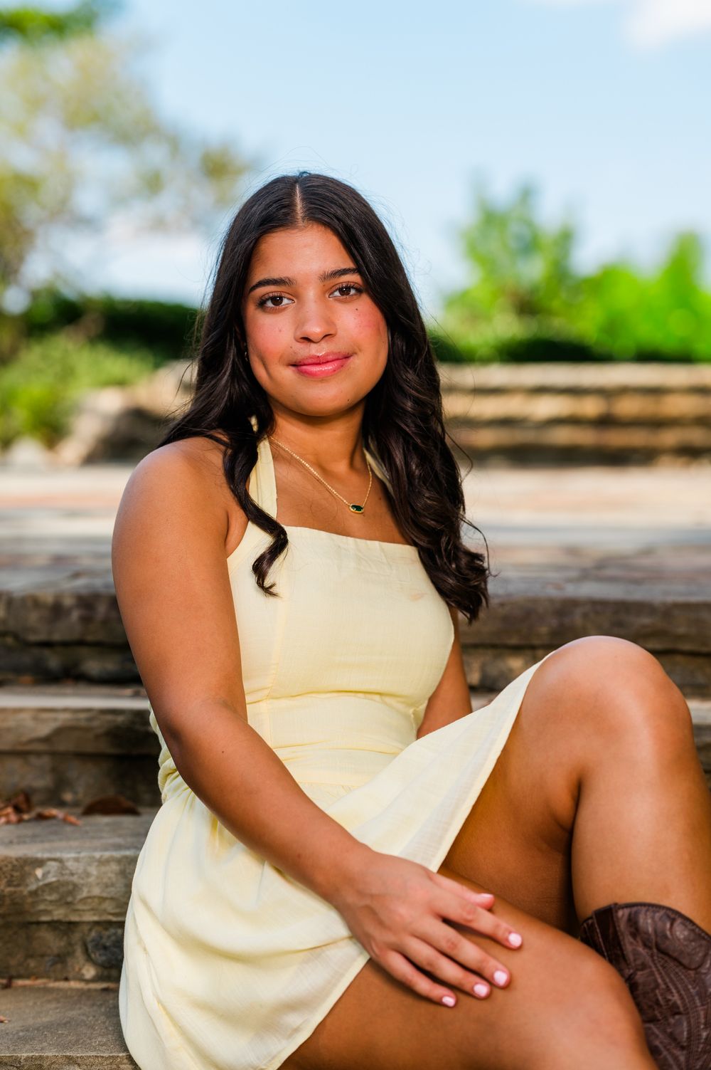 Young woman in yellow dress sitting on stone steps outdoors, smiling at camera with lush greenery background.