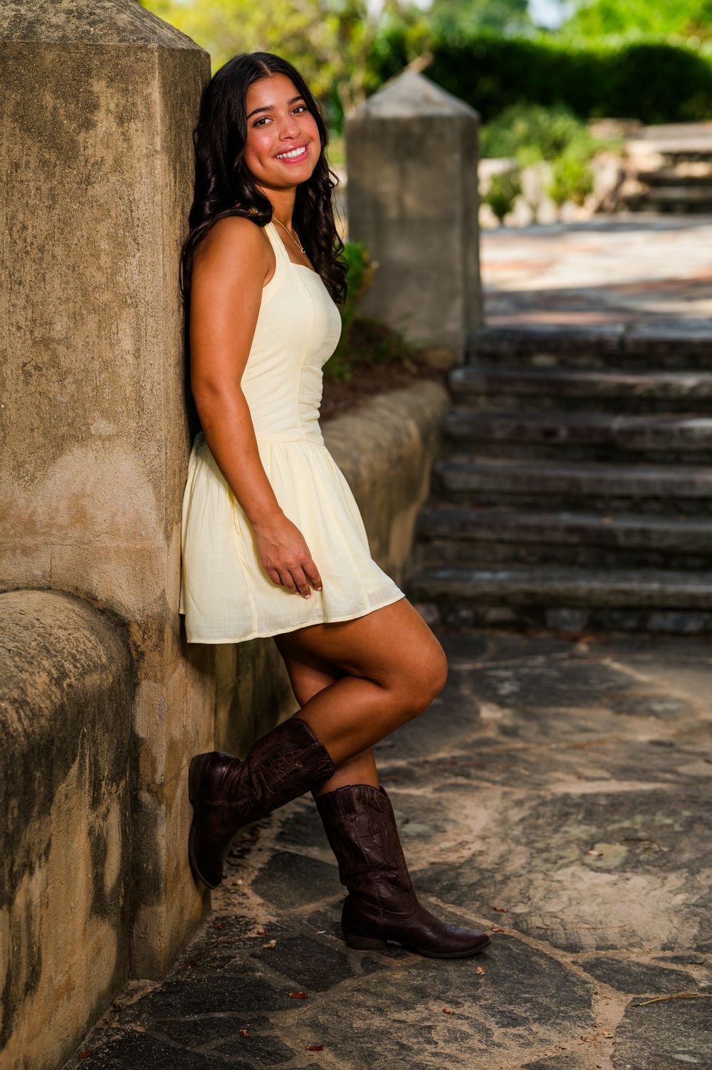 Young woman in white dress and brown cowboy boots leaning against stone wall smiling outdoors.
