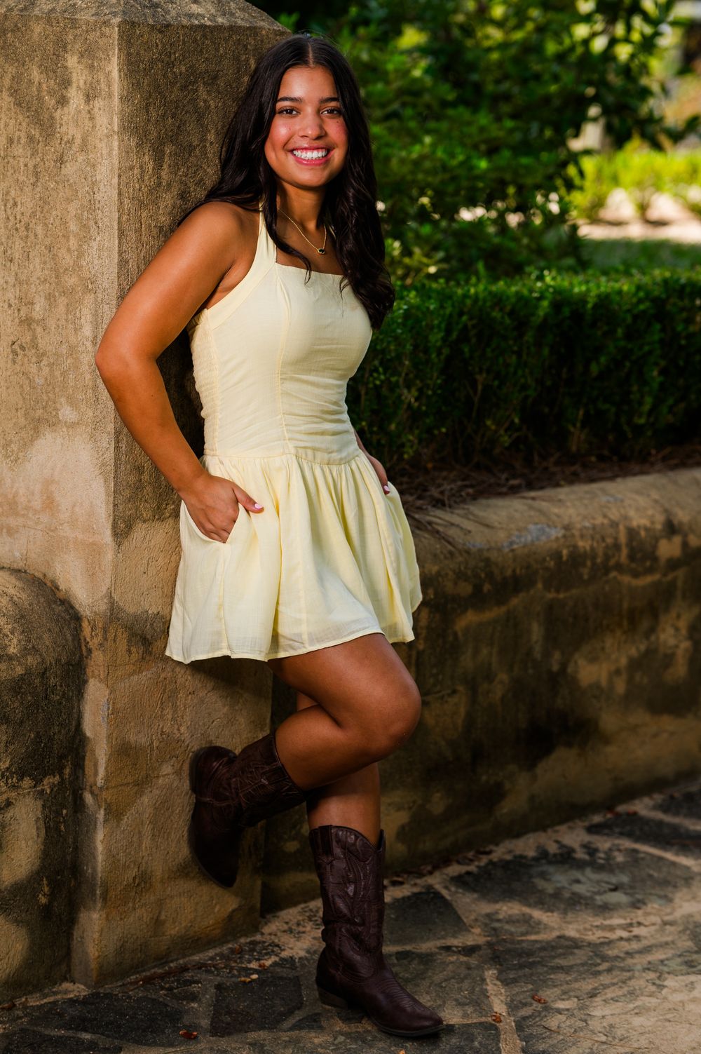Smiling young woman in yellow mini dress and brown cowboy boots leaning against stone wall outdoors.
