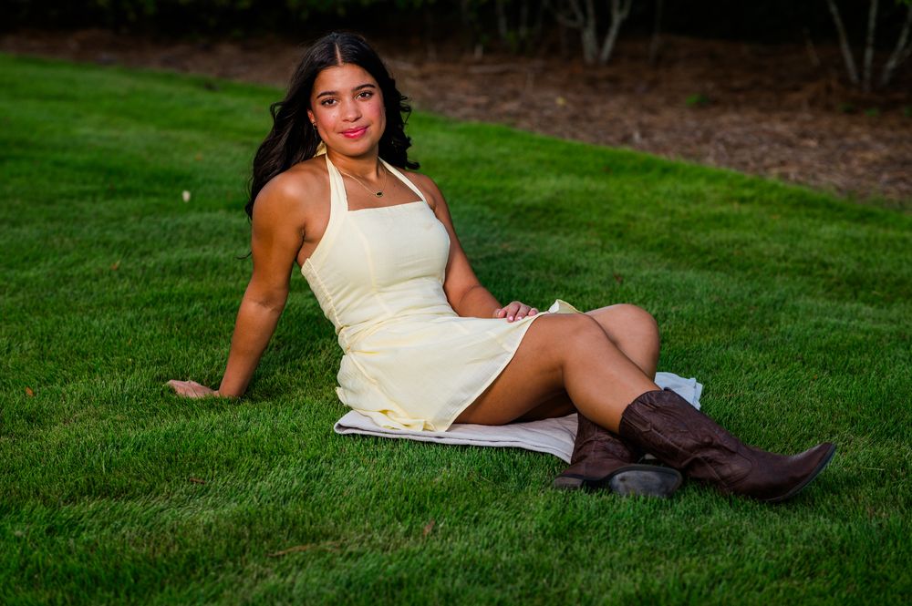 Young woman in white dress and brown cowboy boots sitting on green grass outdoors.