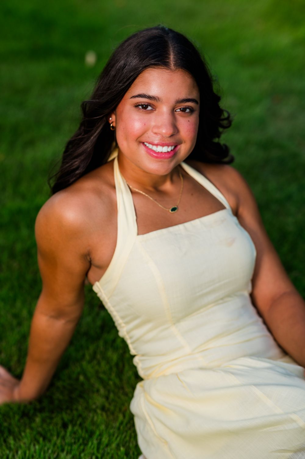 Young woman in cream halter dress sitting on green grass, smiling outdoors during a portrait session.