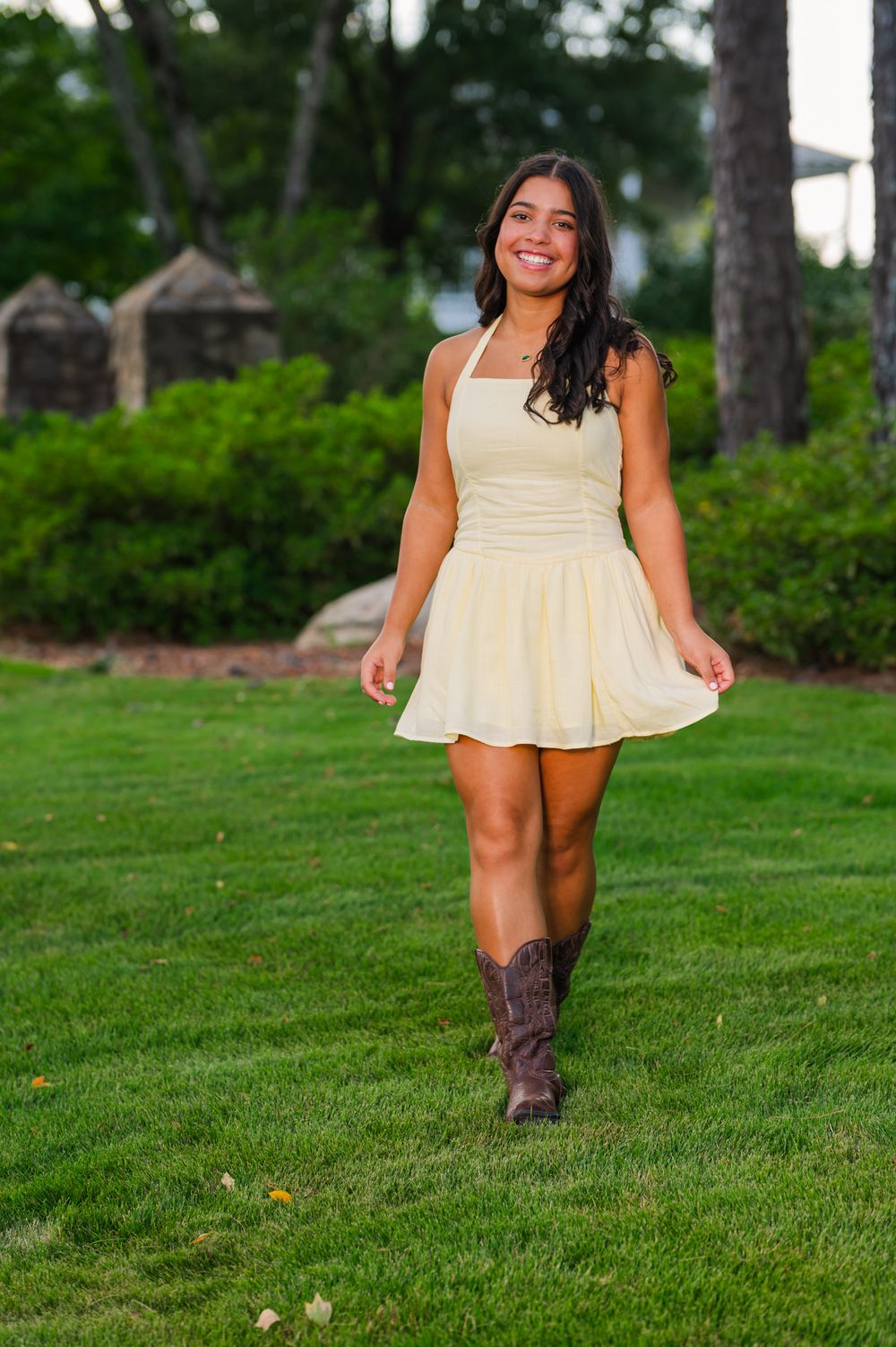Young woman in yellow mini dress and cowboy boots smiling while walking on green grass outdoors.