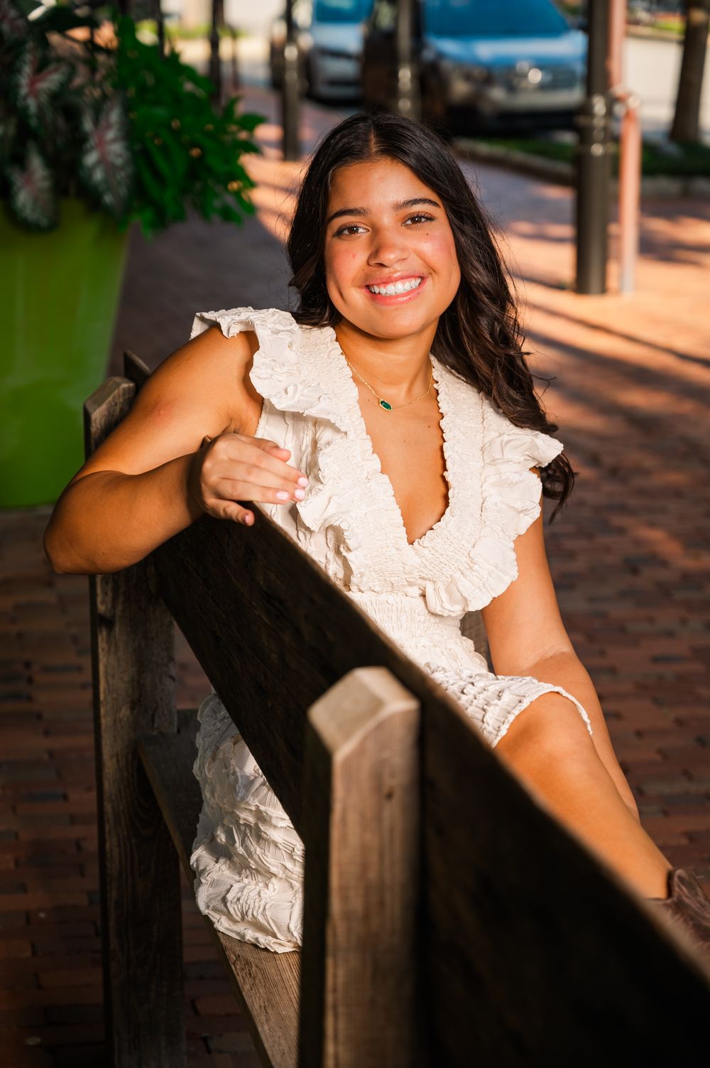 Smiling young woman in white ruffled dress leaning on wooden railing outdoors on brick walkway.