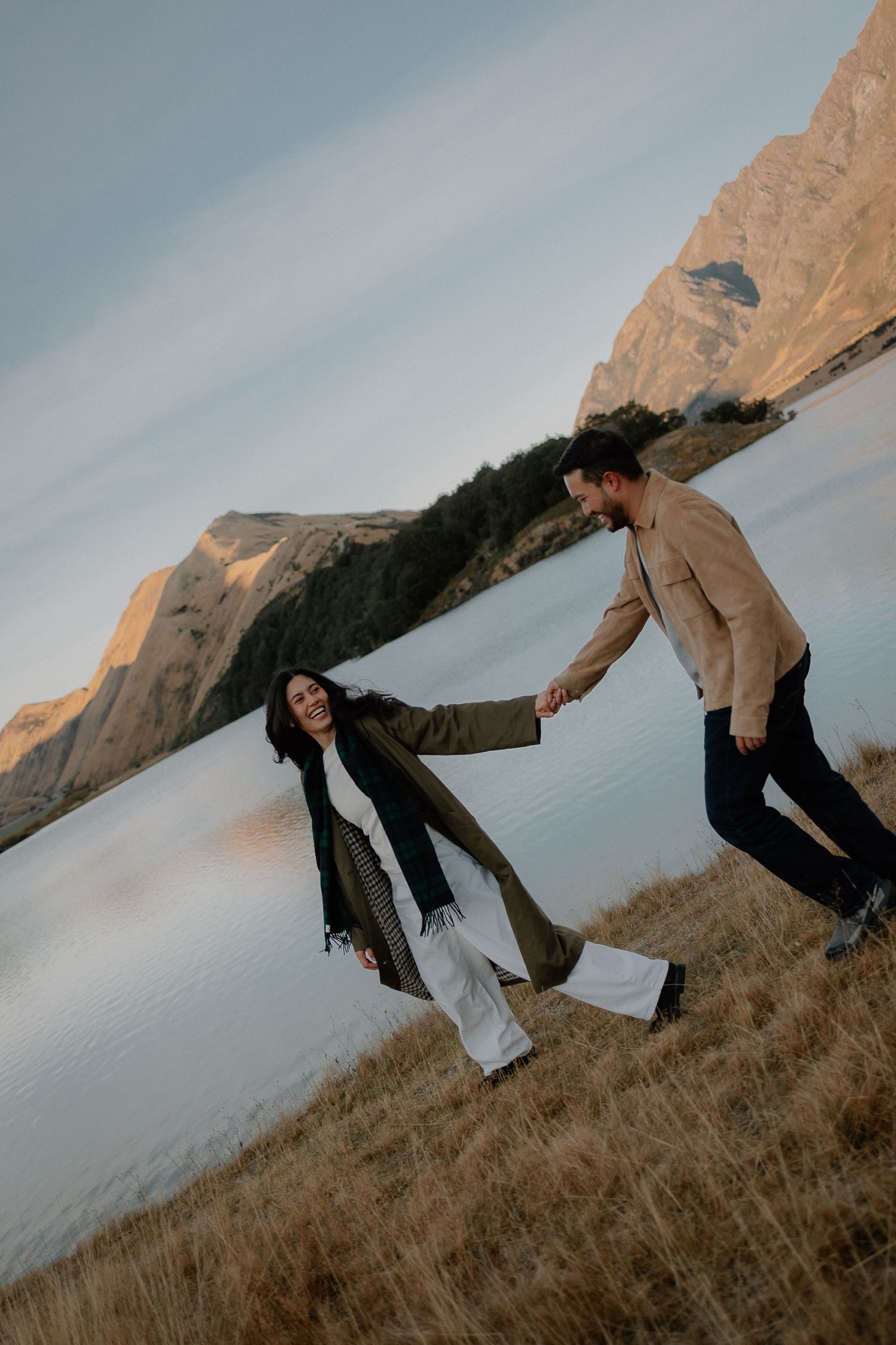 Couple dancing in golden field with mountain lake backdrop in warm sunlight.