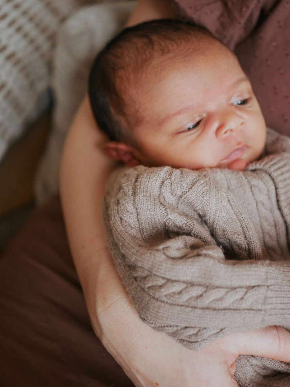 Newborn baby in beige cable-knit sweater resting peacefully in parent's arms.