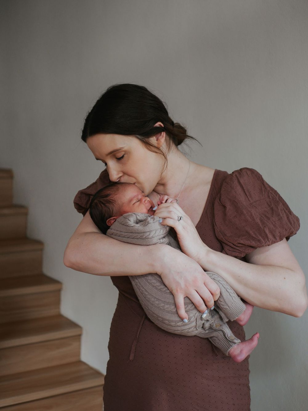 Mother in brown dress kisses her newborn baby while standing near a staircase.