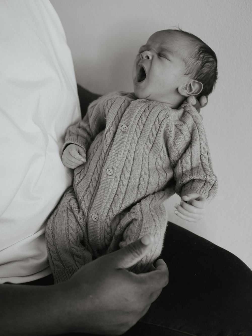 Black and white photo of yawning newborn baby in cable-knit onesie held by parent.