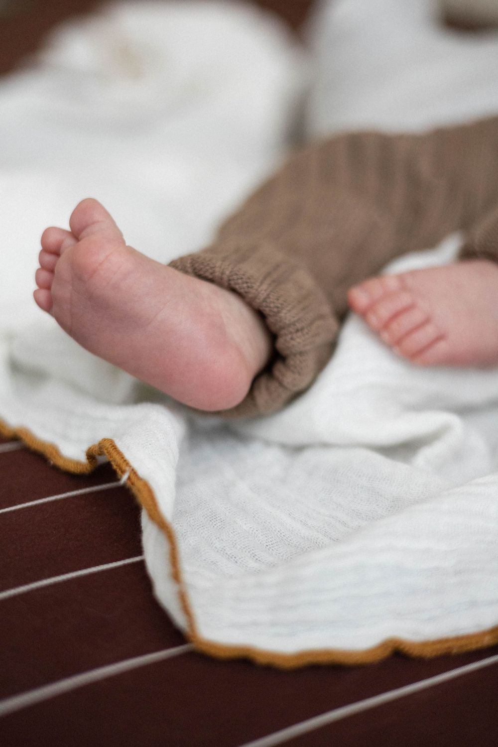 Close-up of tiny newborn baby feet peeking from brown knit pants on white blanket.