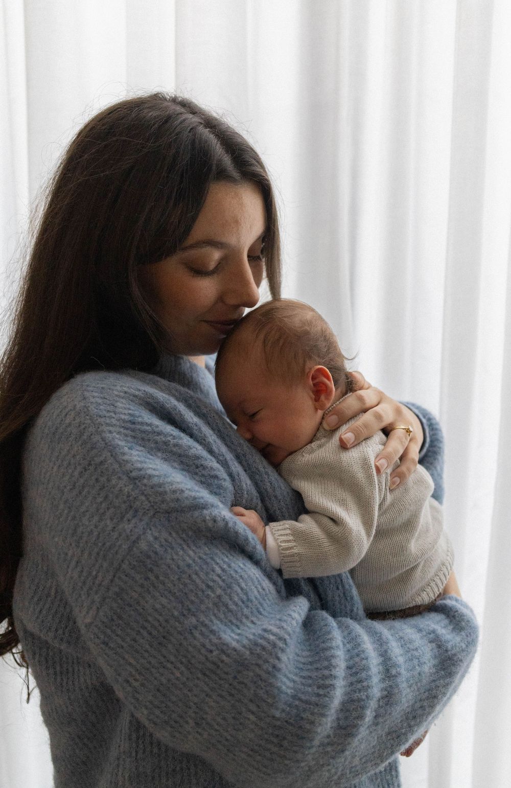 New mother in blue sweater holds and kisses her sleeping newborn baby by a window.