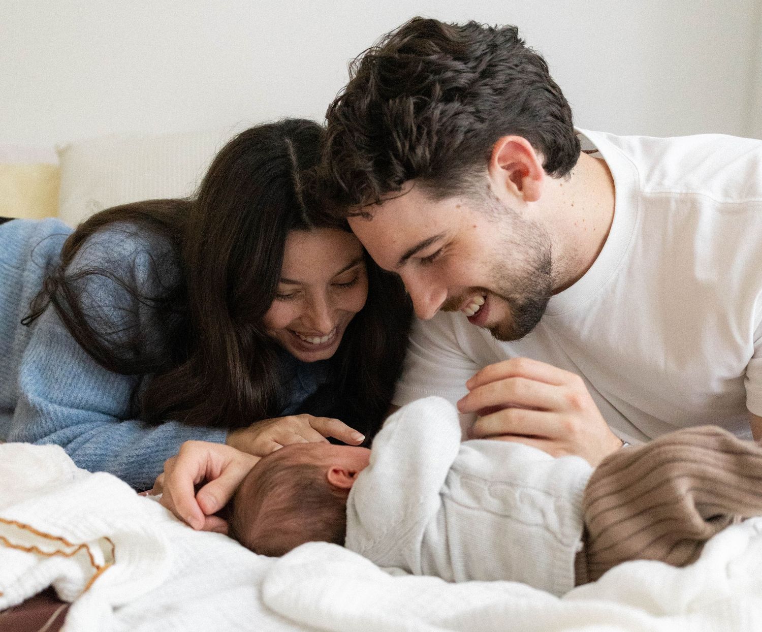 Happy young parents smiling and bonding with their newborn baby lying on a white bed at home.