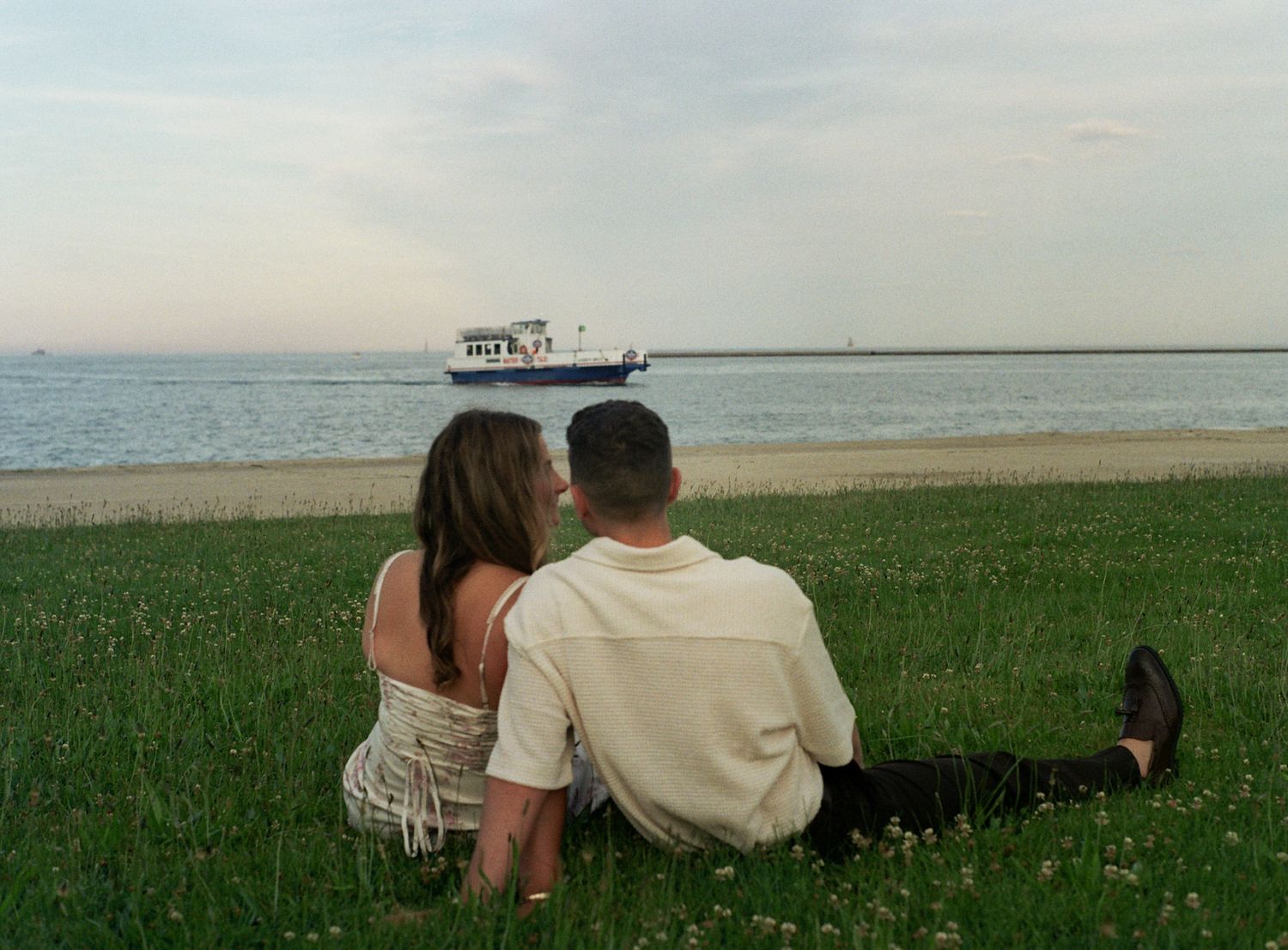 Couple sitting on green grass by the waterfront watching a white ship sailing on calm waters at dusk. shot on 35 mm film