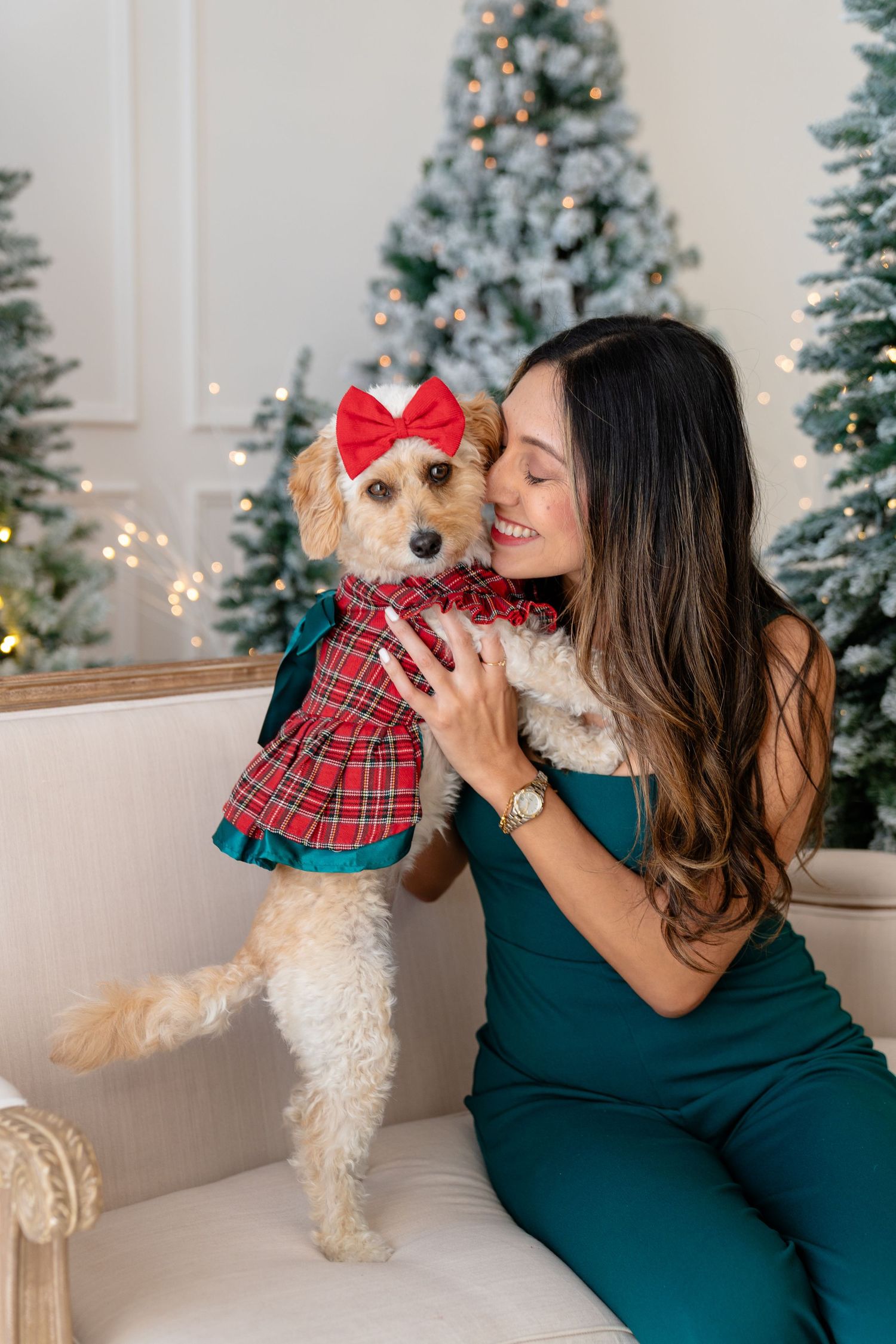 Woman in green jumpsuit holding a dressed Goldendoodle dog with red bow near Christmas trees.