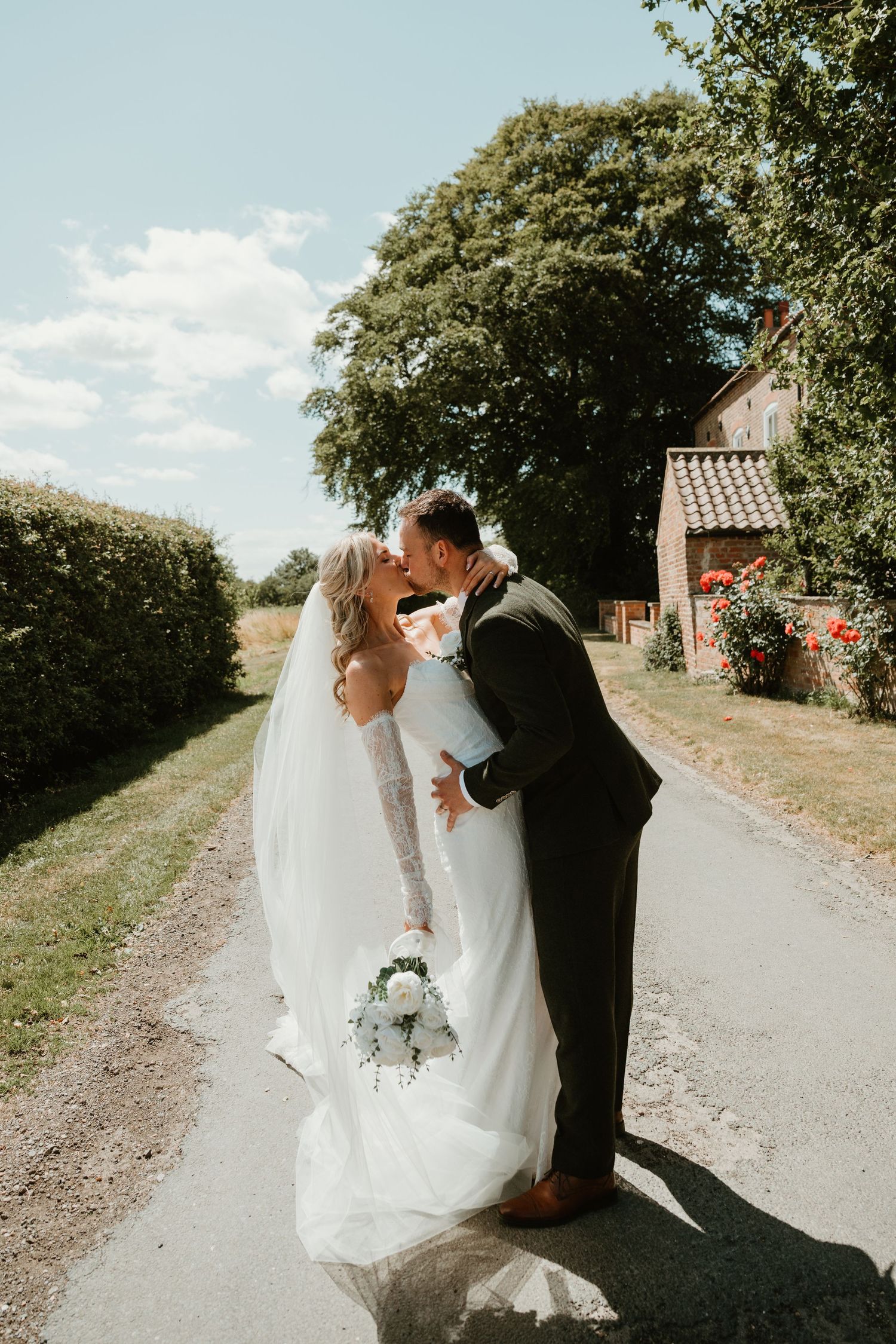 Bride and groom share a romantic kiss on a sunlit country path, bride holding white bouquet in flowing wedding dress.