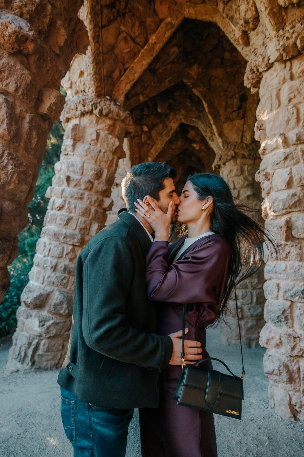 A couple kissing passionately after a proposal, with the golden chariot statues and rushing waterfall of park guell in the background.