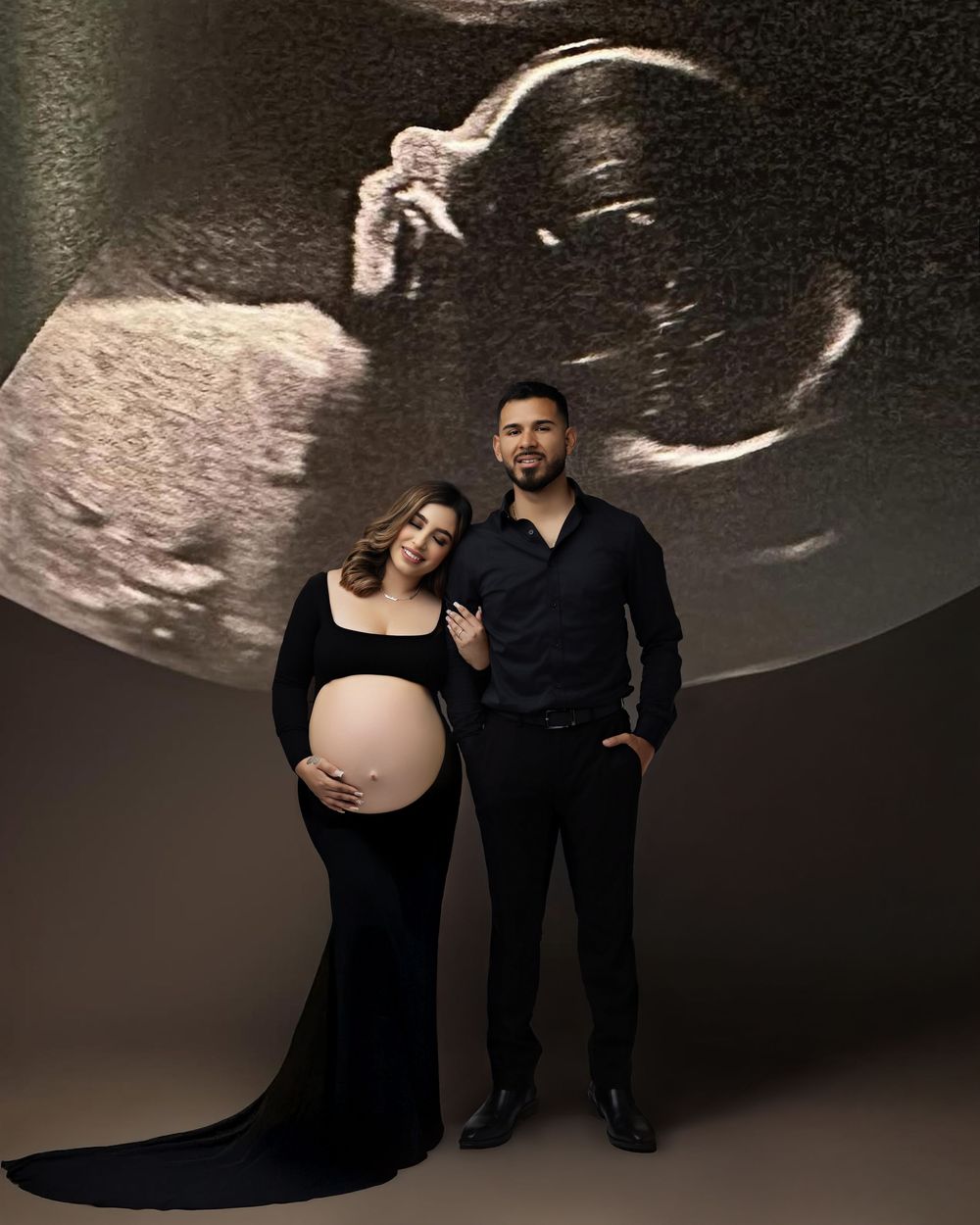 Pregnant couple in black outfits posing for maternity photo with ultrasound image projected behind them.