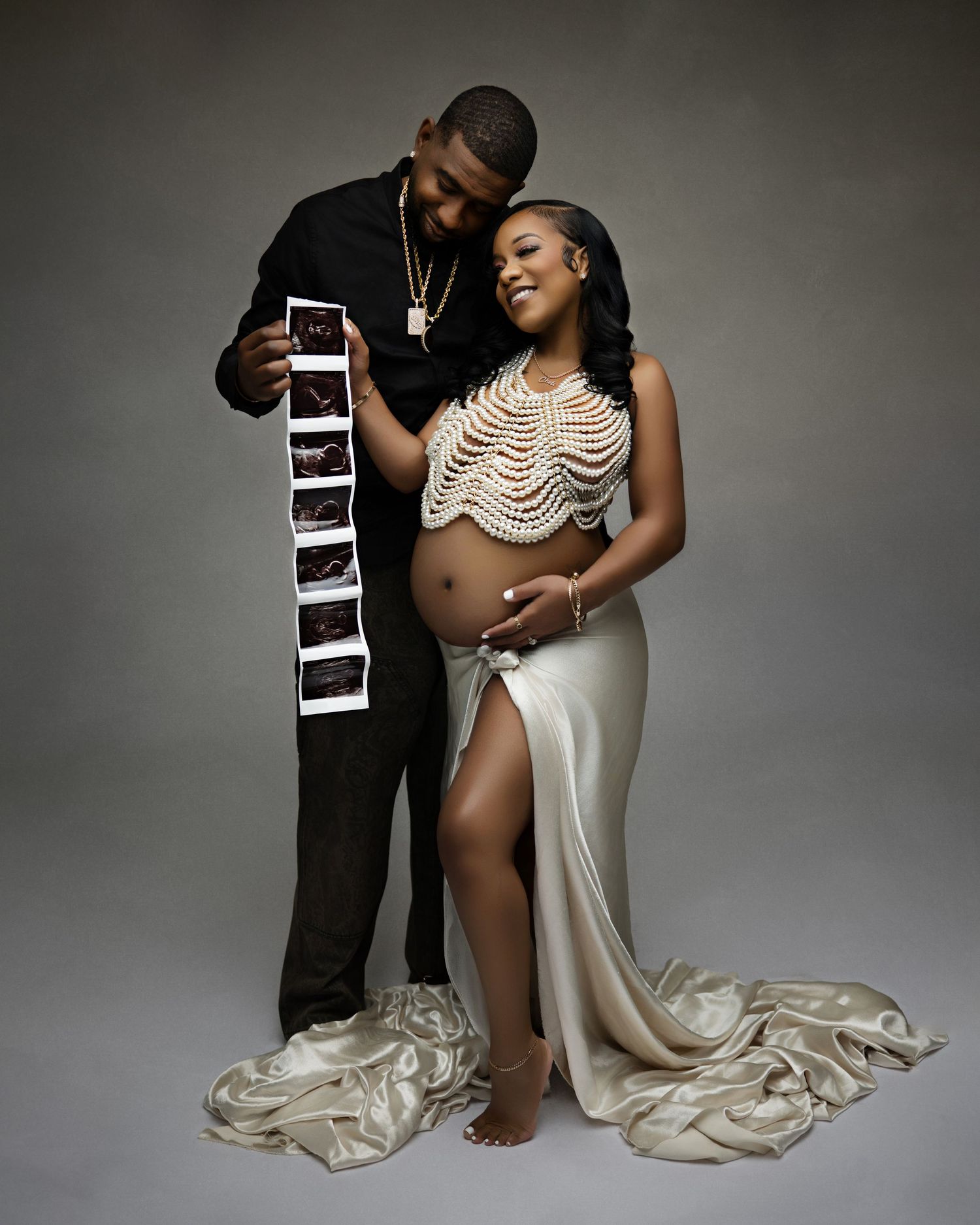 Couple maternity photo with pregnant woman in pearl top and satin skirt, partner holding ultrasound images.