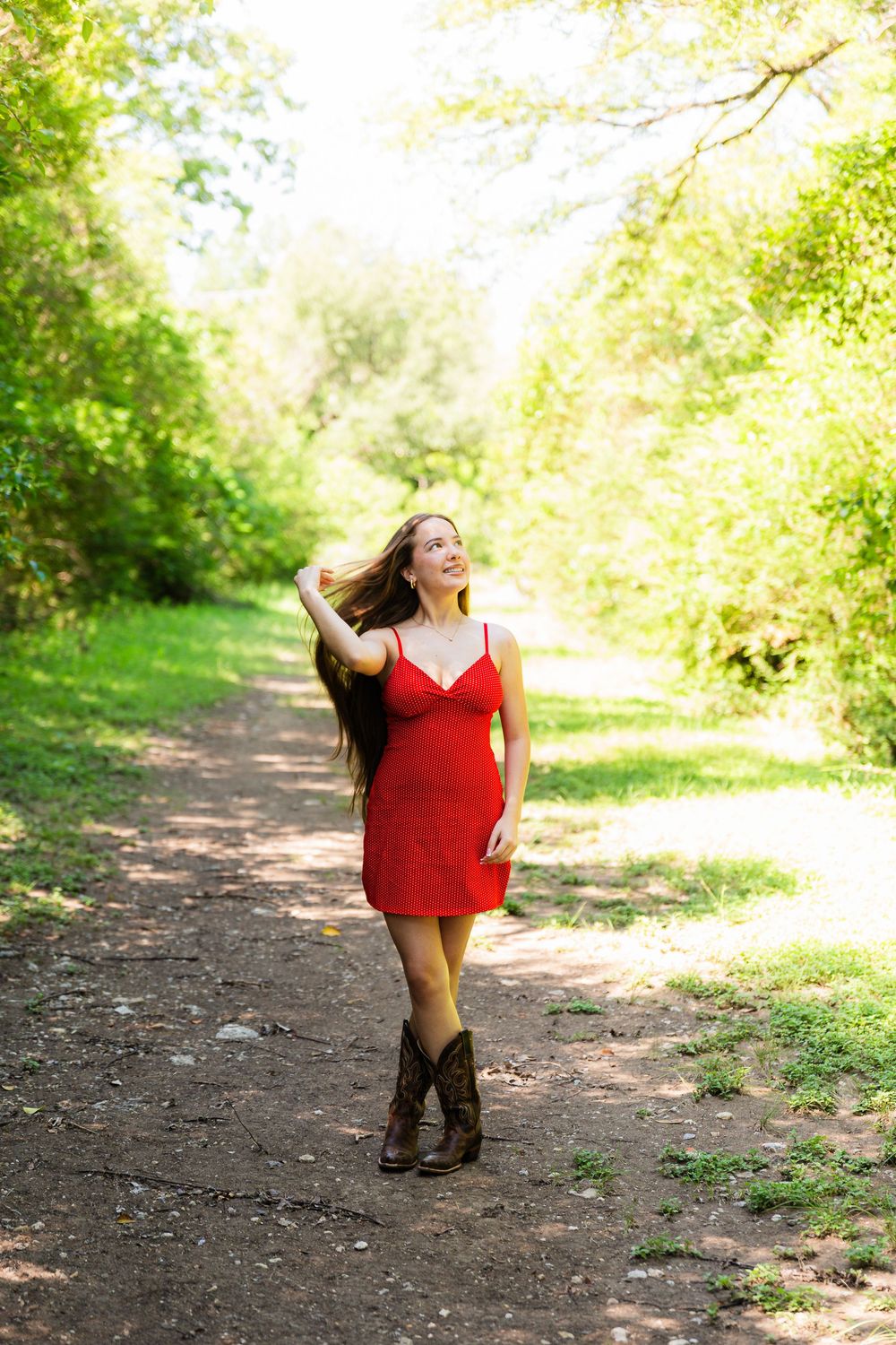 Young woman in red dress and cowboy boots walking on a sunlit forest path.