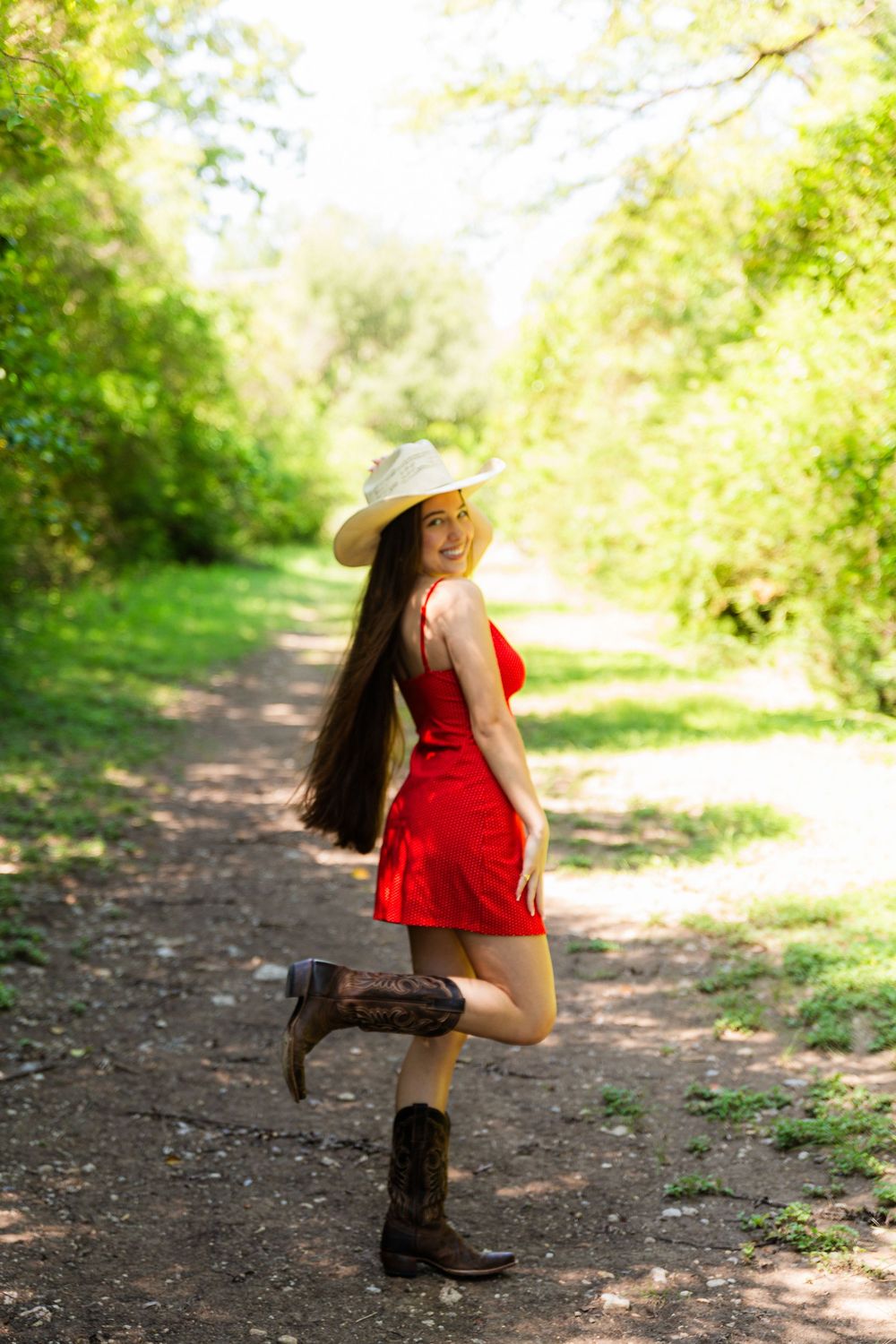 Young woman in red dress, cowboy hat, and boots smiling on a sunny forest path.