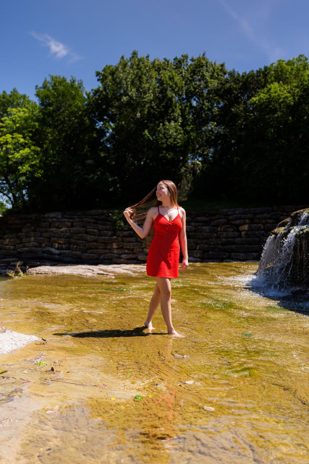 Woman in red dress walking barefoot in shallow water near a waterfall on a sunny day.