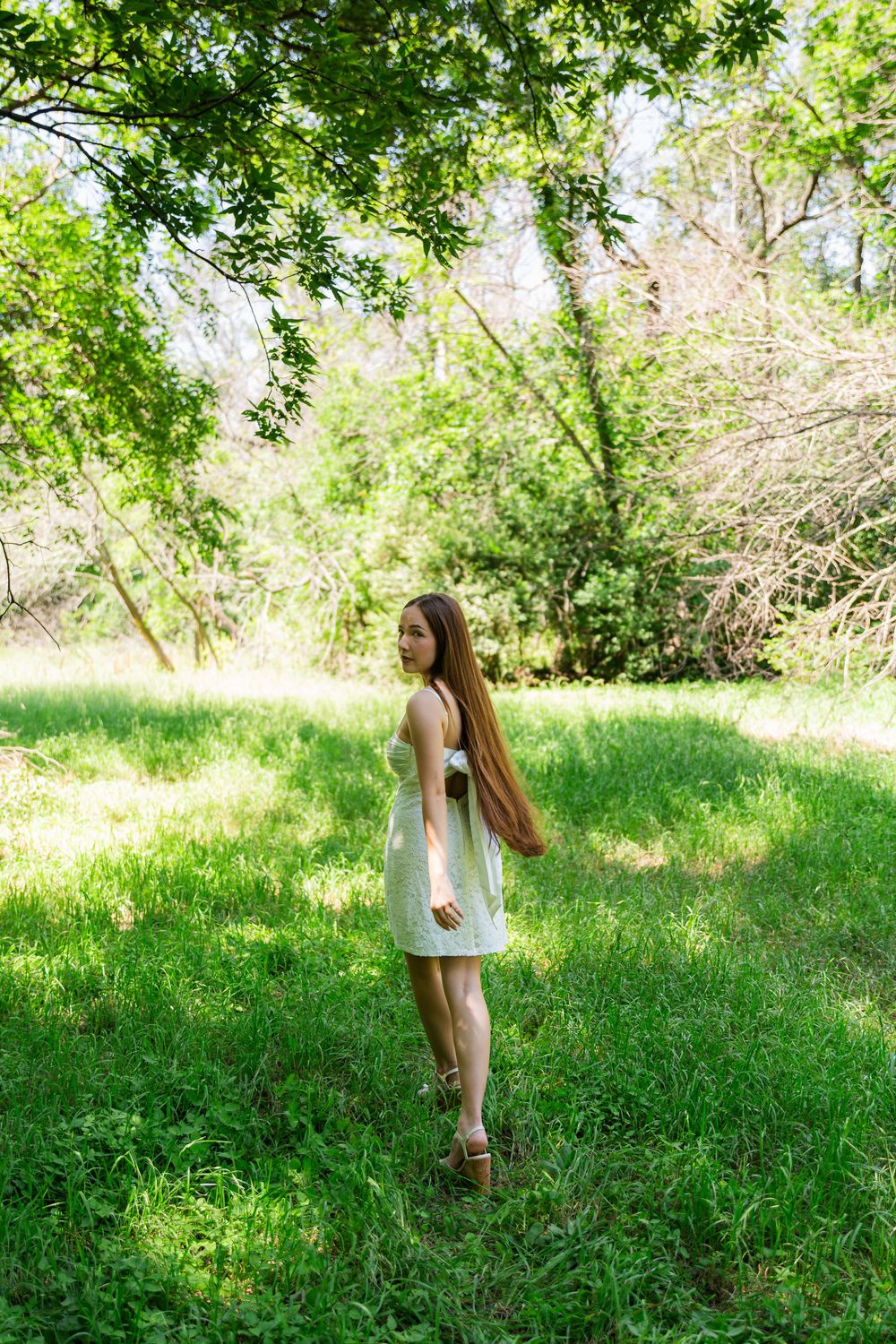 Young woman with long hair in a white sundress standing on green grass in a sunny park.