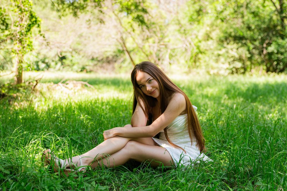 Young woman in white dress sitting on lush green grass in a sunny park, smiling serenely with long brown hair.