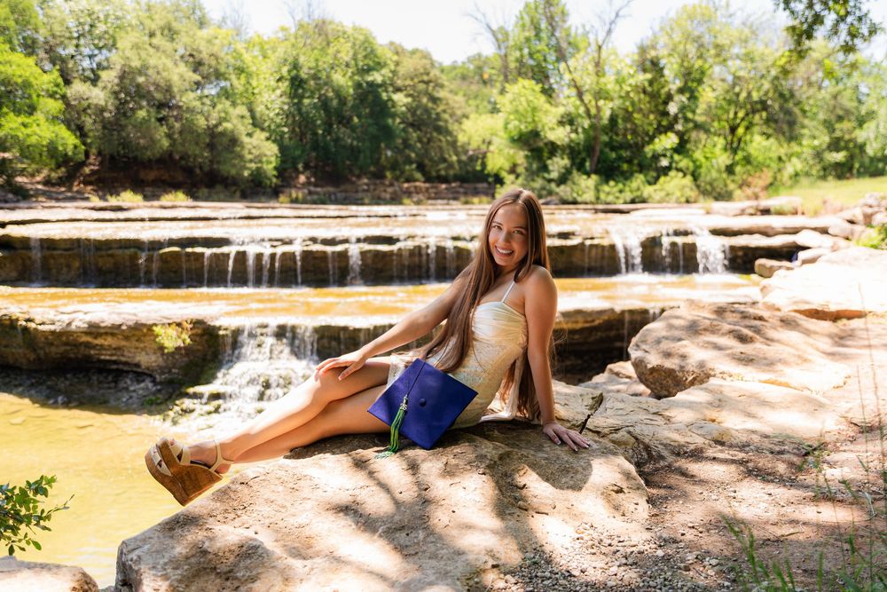 Smiling young woman sitting on rocks beside a scenic waterfall on a bright sunny day.