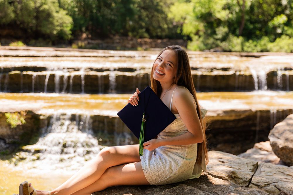 Young woman smiling outdoors, holding a blue book, sitting on rocks near a scenic waterfall in a lush green setting.
