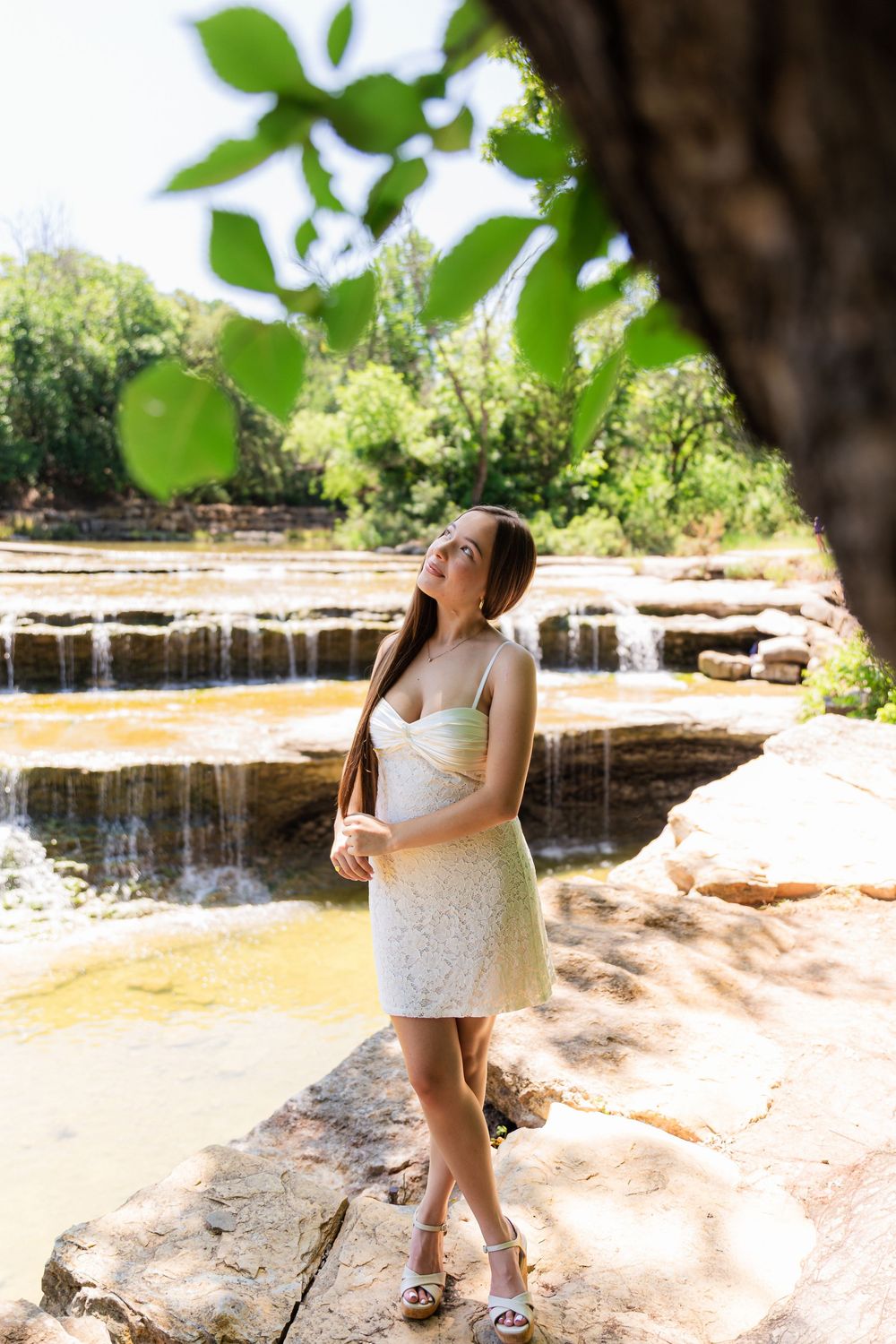Young woman in white lace dress standing on rocks near a cascading waterfall surrounded by lush greenery.