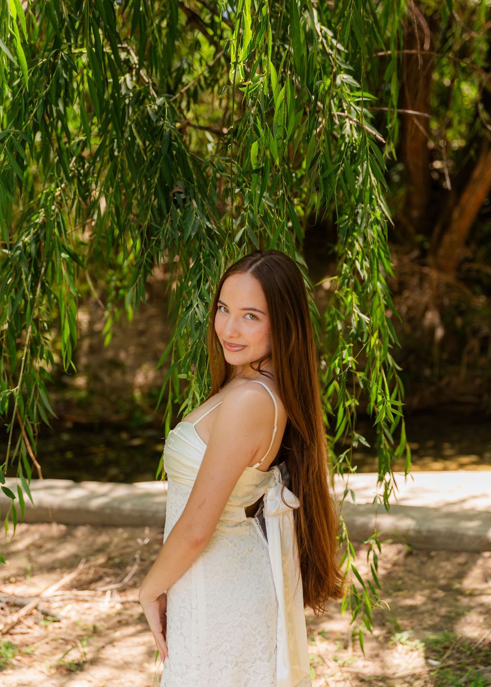 Young woman with long brown hair in white dress poses under weeping willow tree outdoors.