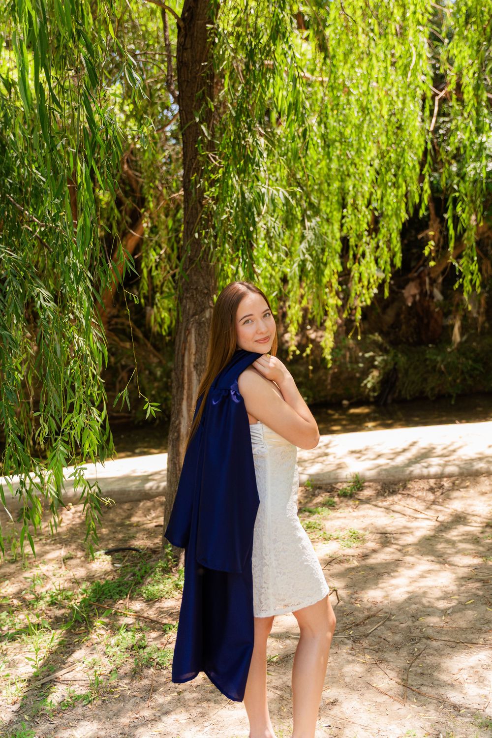 Young woman in white dress holding blue graduation gown under weeping willow tree.